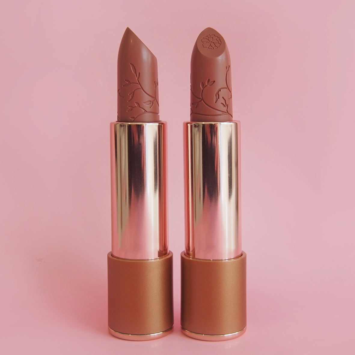 Two nude lipsticks with gold caps on a pink background